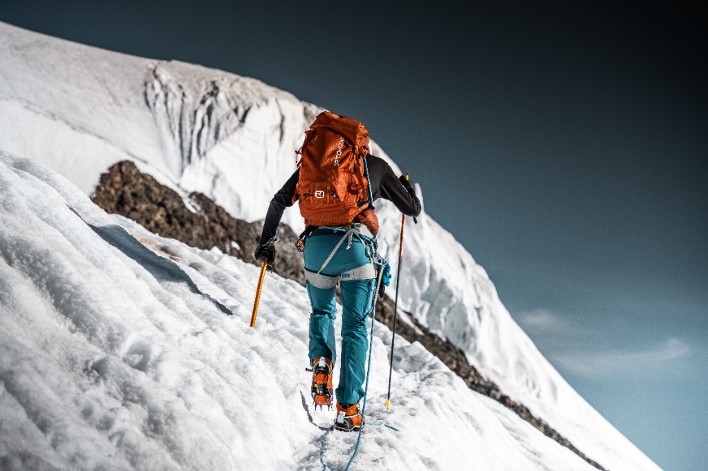 A climber with an orange pack walking up a snowy ridge